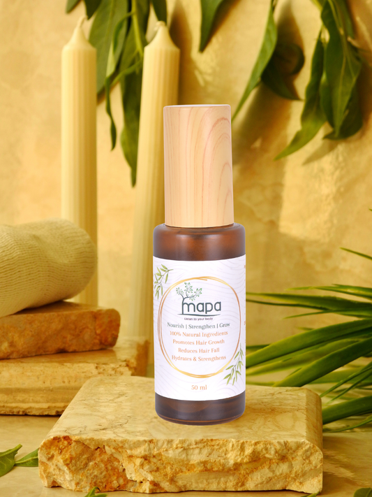 A bottle of mapa Hair Care hair oil stands on a textured stone slab, surrounded by natural props including green leaves, candles, and a rolled towel against a warm beige background.